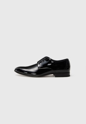Zapato de vestir para hombre de piel negra pulida con cordones y tacón bajo, sobre un fondo gris claro liso.