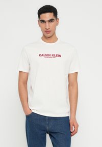 Νεαρός άνδρας φορώντας ένα λευκό μπλουζάκι Calvin Klein με κόκκινο λογότυπο και μπλε τζιν, στέκεται μπροστά σε ένα απλό φωτεινό φόντο.