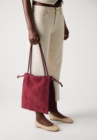 Sac à cordon en suédine bordeaux avec des bretelles, offrant une texture douce et un design minimaliste. Porté avec un pantalon large crème.