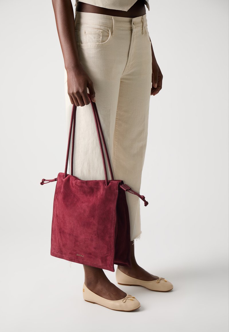 Sac à cordon en suédine bordeaux avec des bretelles, offrant une texture douce et un design minimaliste. Porté avec un pantalon large crème.