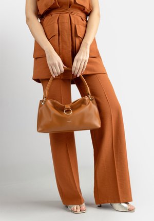 Donna con un abito color ruggine che tiene una borsa in pelle marrone chiaro con dettaglio ad anello argentato, indossando tacchi aperti di colore chiaro su uno sfondo semplice.