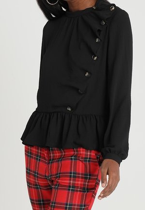 Kvinde iført en sort langærmet bluse med knapper i siden og flæsekant, kombineret med røde og sorte ternede bukser.