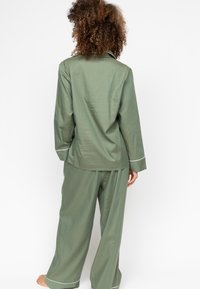 Grünes Pyjama-Set mit einem Hemd mit Kragen und weitgeschnittenen Hosen, das weiße Paspelierungen entlang des Kragens und der Manschetten aufweist. Weicher Stoff.