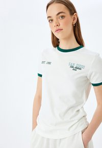 Hvid t-shirt med grøn ribbet krave og ærmekanter. Broderet tekst inkluderer "SAN DIEGO" og "EST. 1992." Blødt bomuldsstof.