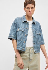 Μπλε υφαντό σακάκι με κοντά μανίκια, εμπρόσθιες τσέπες και λεπτομέρειες με χρυσά κουμπιά· φορεμένο πάνω από λευκό t-shirt και ανοιχτό τζιν παντελόνι.