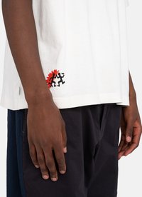 White cotton T-shirt with black and red graphic design featuring figures on the left hem. Short sleeves, relaxed fit, soft texture.
