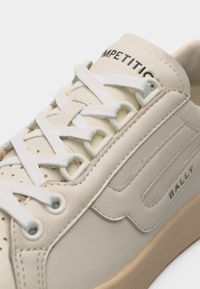 Gros plan sur une basket en cuir beige avec des lacets blancs, des coutures détaillées, la marque "BALLY" sur le côté et l'inscription "COMPETITION" sur la languette.