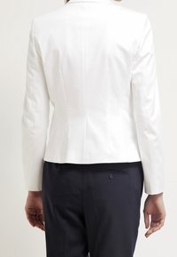 Bílý blazer s přiléhavým střihem, hladkým povrchem a jemnými švy. Kombinován s tmavými kalhotami, které mají zadní kapsu a knoflíkový detail.