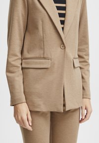 Blazer ajustado de color marrón claro con cierre de un solo botón y bolsillos de parche. Textura suave con un diseño minimalista.