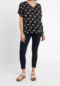 Černá blůza s vzorem plameňáků, krátké rukávy a výstřih do V. Kombinováno s tmavými slim-fit kalhotami a bílými sandály na řemínky.