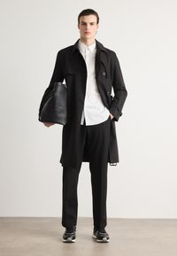Abrigo negro de doble botonadura, camisa blanca debajo, pantalones negros y zapatillas negras. Sosteniendo un bolso negro de hombro.