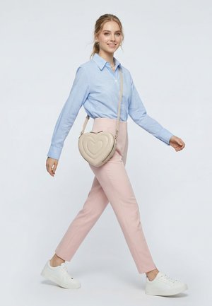 Junge Frau geht spazieren, trägt ein blau kariertes Hemd, pinke Hosen, weiße Turnschuhe und eine beigefarbene, herzförmige Umhängetasche.