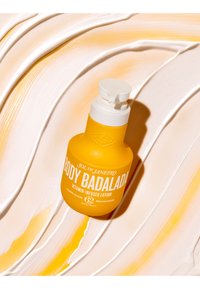 Klar gul Sol de Janeiro Body Badalada lotionflaske med hvit pumpe står på kremet, virvlet lotionbakgrunn.