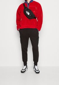 Chaqueta de forro polar roja, cuello azul, pantalones cargo negros con bolsillos y zapatillas negras y blancas. Riñonera con logotipo en la parte frontal.
