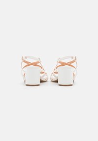 Vita blockklack-sandaler med korsade remmar, med en ljus orange accent och en slät textur. Bakifrån ses ankelremmen med spänne.