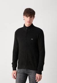 Maglione zip-up nero a coste con colletto alto, dotato di un piccolo logo sul lato sinistro del petto e polsini aderenti. Il materiale appare testurizzato.