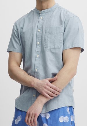 Mężczyzna w jasnoniebieskiej koszuli na krótki rękaw z kieszenią na piersi oraz niebieskich szortach z białymi wzorami muszelek, stojący na jednolitym tle.