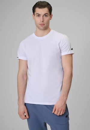 Młody mężczyzna w prostym białym t-shircie z krótkim rękawem i niebieskich spodniach dresowych stoi na tle szarego tła.