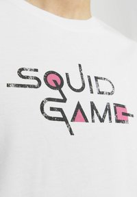 Hvid bomulds t-shirt med teksten "SQUID GAME" i slidte sorte og pink farver samt geometriske detaljer, rund halsudskæring, korteærmer.