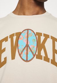 Beige tričko s nápisem "EVOKE" hnědými písmeny. Obsahuje design symbolu míru v modré barvě s květinovými vzory, obšitý hnědou výšivkou.