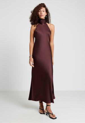 Donna con capelli ricci che indossa un abito senza maniche in raso bordeaux scuro con collo ad anello e sandali neri con tacco a listini, in piedi su pavimento grigio.