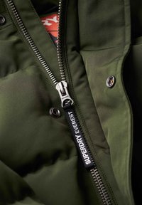 Zielona kurtka puchowa z srebrnym zamkiem i czarnym uchwytem z logo. Posiada pikowaną teksturę oraz zatrzaskowy detal przy kołnierzu.