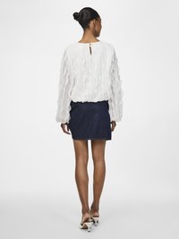 Blusa bianca e strutturata con maniche a volant, abbinata a una gonna di jeans scura. Indossata con mule bianchi con tacco, dettaglio a chiave sulla schiena visibile.