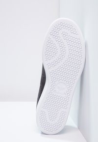 Sole černých tenisek Adidas s bílým hexagonálním vzorem a logem Adidas, stojícími vzpřímeně proti bílé zdi.