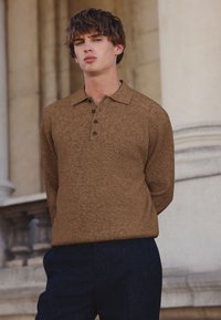 Brauner gerippter Pullover mit Kragen und einer Knopfleiste mit drei Knöpfen, kombiniert mit dunkelblauen Hosen. Strukturiertes Gewebe.