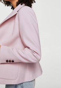 Veste rose clair cintrée avec revers prononcés, tissu texturé et trois boutons foncés en décoration sur le poignet. Ourlet légèrement évasé.