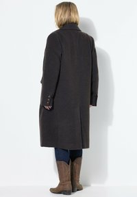 Lange bruine wollen jas met een gestructureerd visgraatpatroon, voorzien van een rugventilatie en knoopmanchetten. Gecombineerd met blauwe jeans en bruine laarzen.