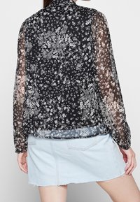 Schwarze Bluse mit floralem Muster und durchsichtigen langen Ärmeln, die weiße Blumenmuster zeigt. Der Saum ist über einen hellen Jeansrock geschichtet.