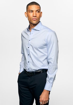 Mann med kort hår og ørepynt, iført en lyseblå skjorte og svarte bukser, ser til siden med hendene i lommene.