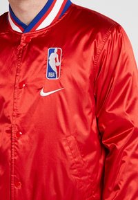 Rote Satin-NBA-Jacke mit Druckknopfverschluss, ausgestattet mit einem weißen Nike-Logo und einem blau-weißen, gestreiften Rippkragen.