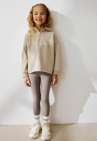 Beige zip-up hoodie med en logotyp, ljusgrå leggings och krämfärgade sneakers med vita och grå detaljer. Släta texturer, avslappnad stil.