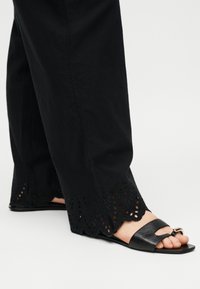 Πόδια με μαύρα σανδάλια ανοιχτών δαχτύλων με χρυσή λεπτομέρεια αγκράφας, σε συνδυασμό με μαύρο παντελόνι με δαντέλα με τρύπες στη γέφυρα.