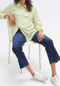 Personne assise sur une chaise blanche portant une chemise à motif vert clair, un jean bleu et des baskets blanches avec des semelles vert clair.