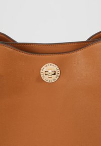 Sac à main en cuir fauve à texture grainée. Il est doté d'un fermoir circulaire doré avec logo, d'une large ouverture et de bords lisses.