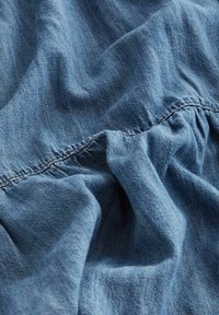 Sininen farkkukangasta, jossa näkyvät saumat ja laskokset luovat tekstuuria ja varjoja pinnalle.