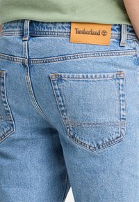 Los pantalones vaqueros de denim azul claro presentan un parche de cuero con el logo de Timberland, costuras visibles y dos bolsillos traseros con diseños angulares.