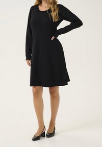 Robe noire à manches longues avec un corsage ajusté et une jupe évasée. Tissu doux, encolure ronde et coutures minimales. Portée avec des escarpins noirs brillants.