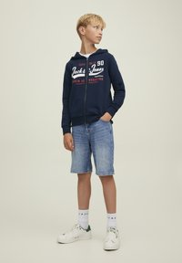 Felpa con zip navy con marchio bianco e rosso, abbinata a pantaloni shorts in denim blu chiaro. Sneakers con dettagli verdi e calzini bianchi.