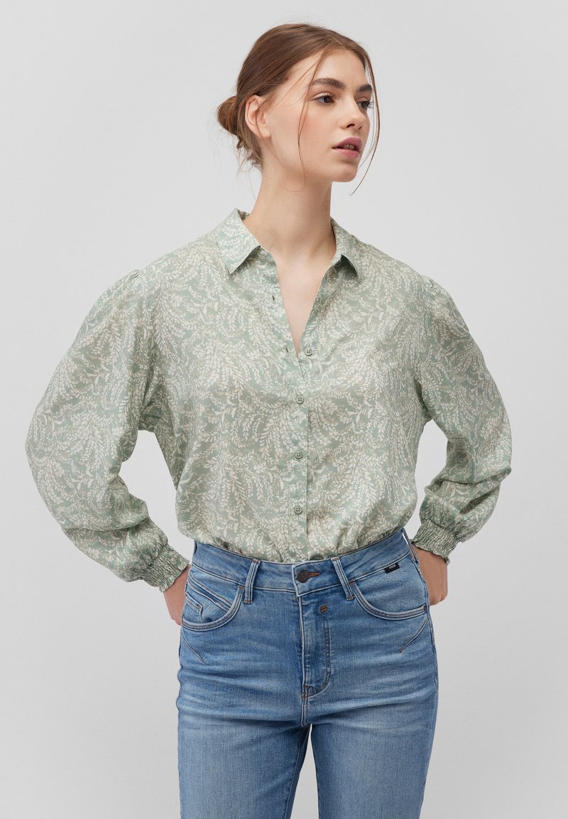 Light green button-up shirt with a floral pattern, long puff sleeves, and elastic cuffs, paired with blue denim jeans.