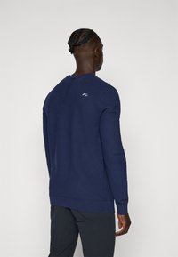 Sweater pullover azul marinho com gola redonda, punhos e bainha canelados, e um pequeno logótipo branco nas costas. Tecido macio e texturizado.