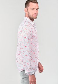 Witte shirt met een roze flamingo patroon, lange mouwen en een lichtblauwe met patroon afgewerkte manchette. De stof lijkt licht en ademend te zijn.