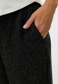 Črne hlače iz teksturirane tkanine z belim pikčastim vzorcem. Roka je postavljena v žep, kar prikazuje gladko, priložnostno obliko in sproščen kroj.