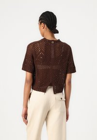 Maglione corto a maniche corte in maglia marrone con orlatura aperta, dotato di uno spacco posteriore e di un collo rotondo. Accompagnato da pantaloni color crema.