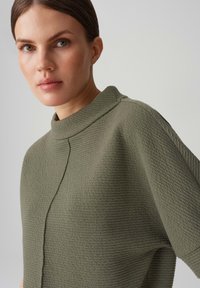 Top verde oliva, strutturato, con colletto alto e maniche corte. Presenta un sottile dettaglio delle cuciture lungo la parte anteriore, realizzato in materiale leggero.