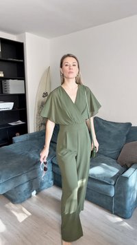 Γυναίκα σε ελαιογράσο jumpsuit κρατώντας γυαλιά ηλίου και τσάντα, στέκεται σε σαλόνι με μπλε καναπέ και σανίδα του σερφ στο φόντο.