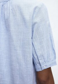 Chemise à manches courtes à carreaux bleu clair et blanc avec poignet boutonné portée sur le bras et l'épaule d'une personne sur fond blanc.